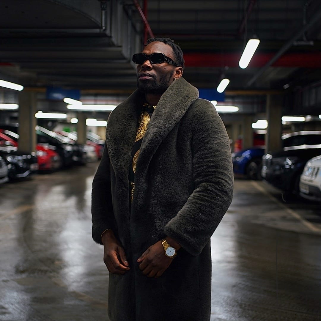 JR in a fur coat and sunglasses standing in a parking garage